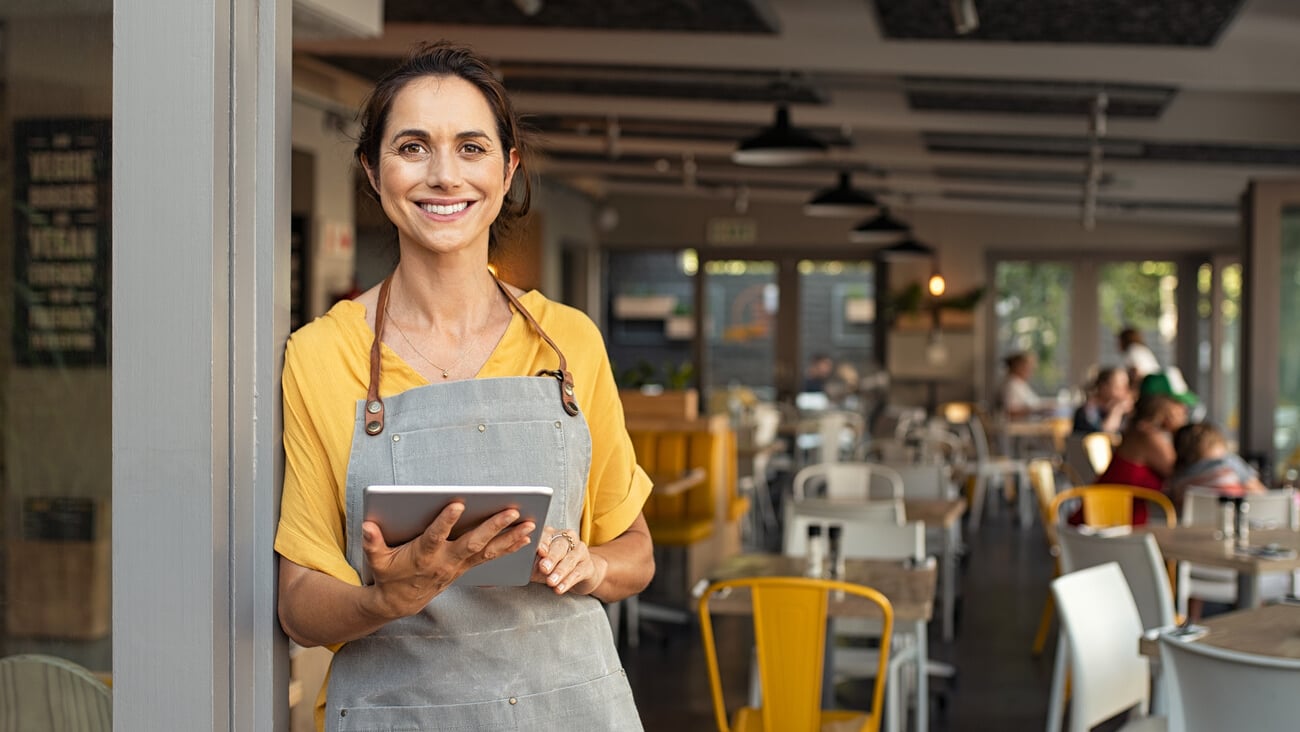 Portrait of happy woman standing at doorway of her store. Cheerful mature waitress waiting for clients at coffee shop. Successful small business owner in casual wearing grey apron standing at entrance; Shutterstock ID 1574160505