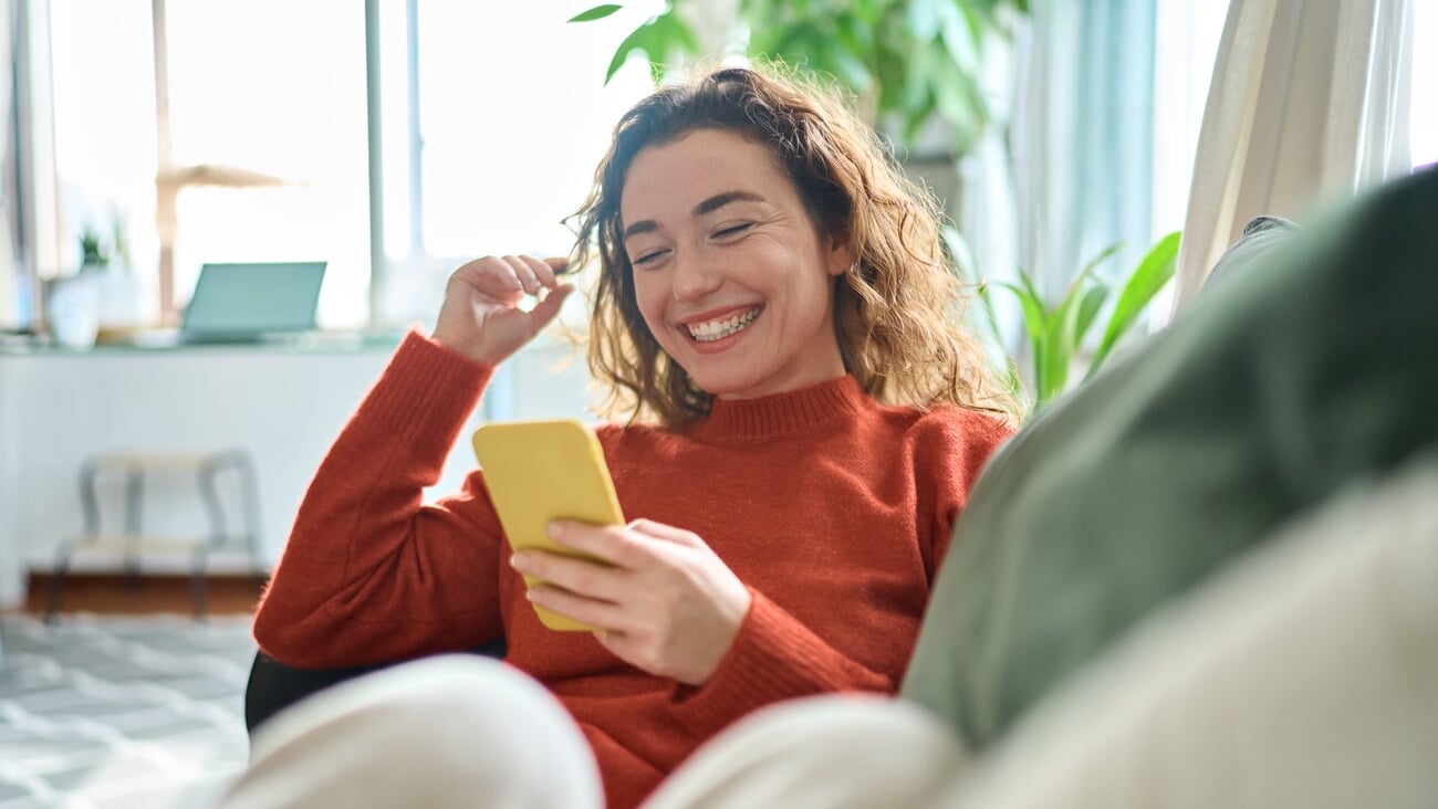 Happy relaxed young woman sitting on couch using cell phone, smiling lady laughing holding smartphone, looking at cellphone enjoying doing online ecommerce shopping in mobile apps or watching videos.; Shutterstock ID 2228645295