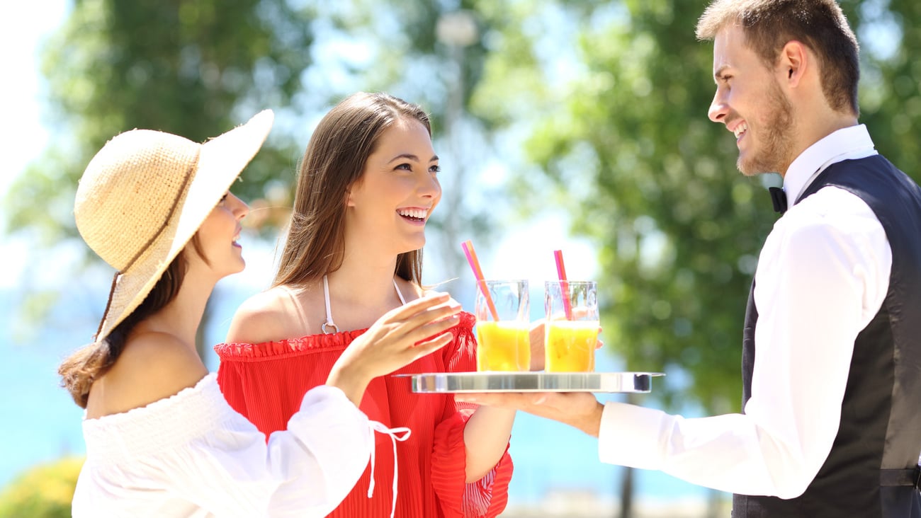 Two hotel customers on summer vacations and a waiter serving them drinks with the ocean in the background; Shutterstock ID 443200663