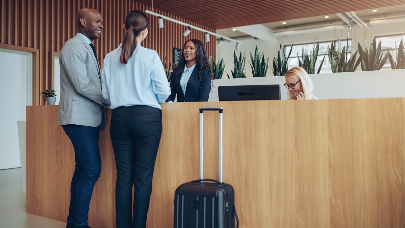 Two smiling guests talking with a concierge while checking in together at the reception counter of a hotel; Shutterstock ID 1477775297