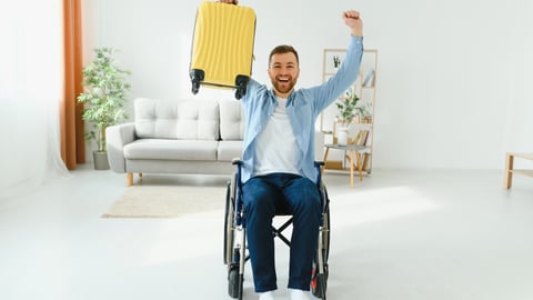 Man in wheelchair holding up yellow suitcase excited for vacation