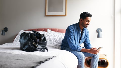 Male guest in boutique hotel sitting on edge of bed checking emails and social media on mobile phone in the morning; Shutterstock ID 1917092945