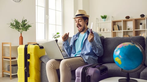 Happy man traveler sitting on home sofa with packed luggage suitcase rejoice successful online ticket booking or hotel reservation holiday vacation with sale discount via laptop computer; Shutterstock ID 1959769993