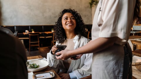 Young smiling african woman using smartphone while paying restaurant bill with contactless payment during dinner party with friends; Shutterstock ID 2322856651