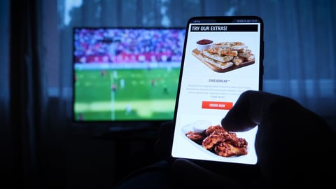 Riga, Latvia - 02/02/2020 EDITORIAL: Background of the TV at home (at the bar), the guy orders different snacks, pizza and sweets scrolling through internet stores using mobile phones. Concept: Sport.; Shutterstock ID 1651678237