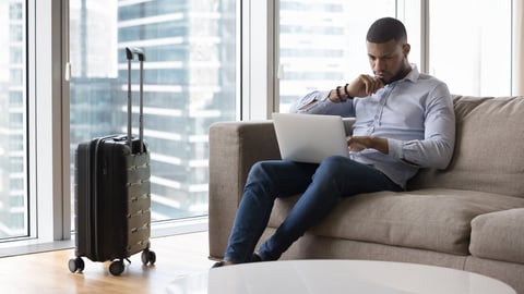 Serious pensive travelling freelance business man using laptop on sofa, working from hotel room, sitting on couch at suitcase, touching chin, thinking over project tasks, looking at monitor; Shutterstock ID 2258228777