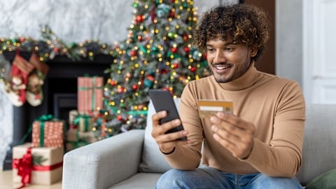 Young man on Christmas at home in the living room sitting near the Christmas tree decorated with Christmas decorations, hispanic smiling holding a bank credit card and phone, choosing gifts online.; Shutterstock ID 2374910895