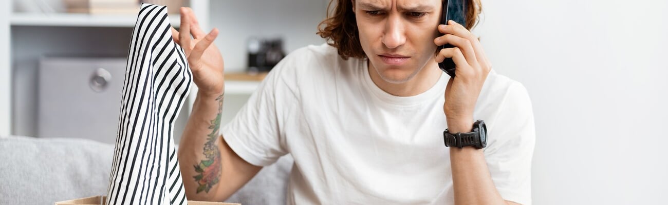 Young Man Unpacking Online Shopping Item, Looks Confused While Talking On Phone, Problem With Order, Suburban Home Background. Indoor, Daylight, Customer Service Issue, Consumer Rights.; Shutterstock ID 2460102123