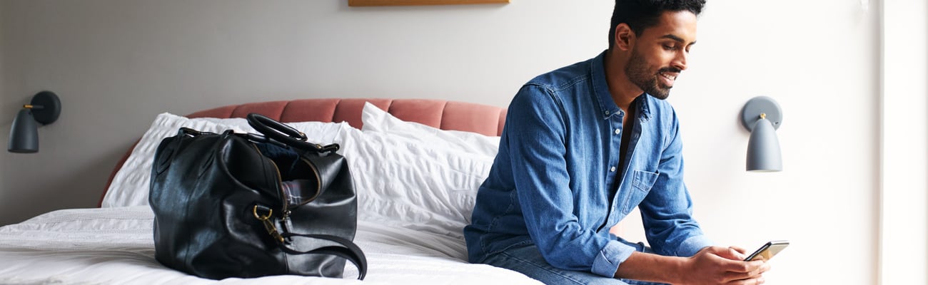 Male guest in boutique hotel sitting on edge of bed checking emails and social media on mobile phone in the morning; Shutterstock ID 1917092945