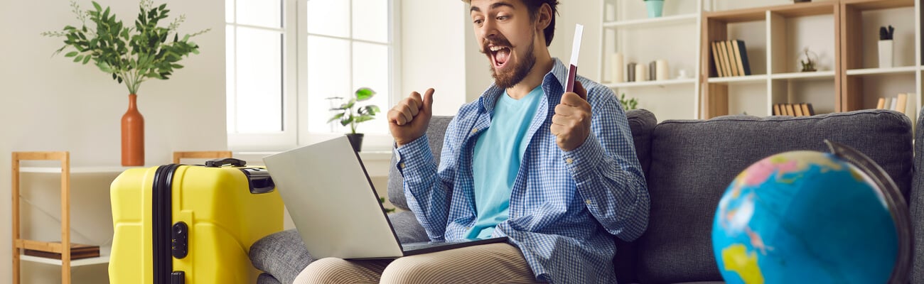 Happy man traveler sitting on home sofa with packed luggage suitcase rejoice successful online ticket booking or hotel reservation holiday vacation with sale discount via laptop computer; Shutterstock ID 1959769993