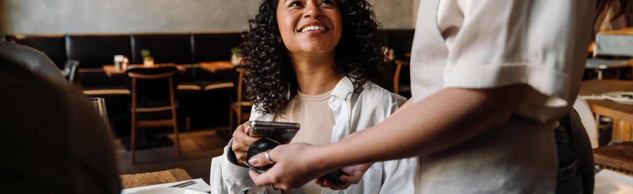 Young smiling african woman using smartphone while paying restaurant bill with contactless payment during dinner party with friends; Shutterstock ID 2322856651