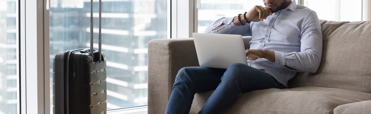 Serious pensive travelling freelance business man using laptop on sofa, working from hotel room, sitting on couch at suitcase, touching chin, thinking over project tasks, looking at monitor; Shutterstock ID 2258228777