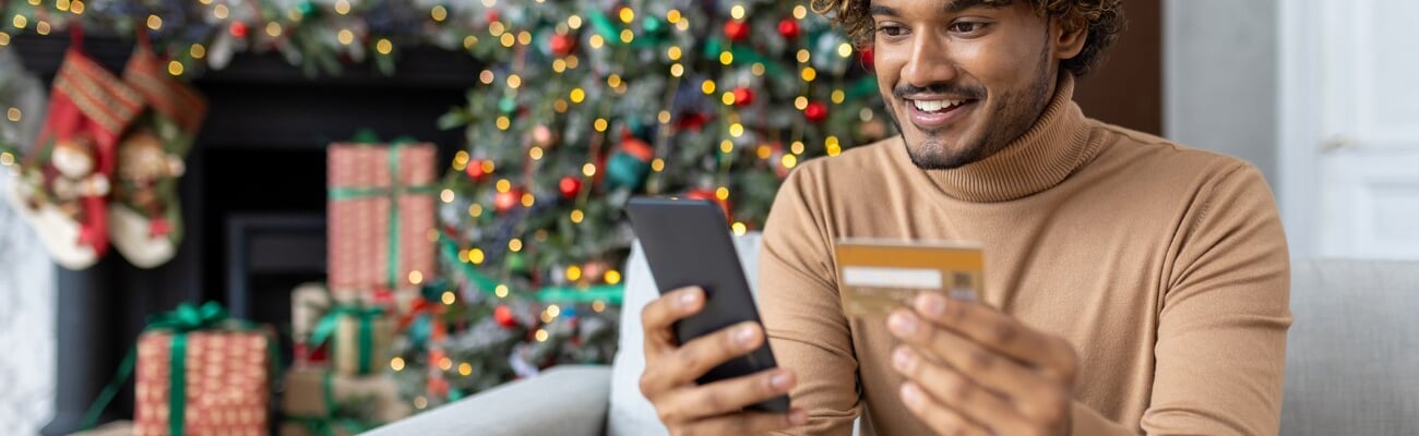 Young man on Christmas at home in the living room sitting near the Christmas tree decorated with Christmas decorations, hispanic smiling holding a bank credit card and phone, choosing gifts online.; Shutterstock ID 2374910895